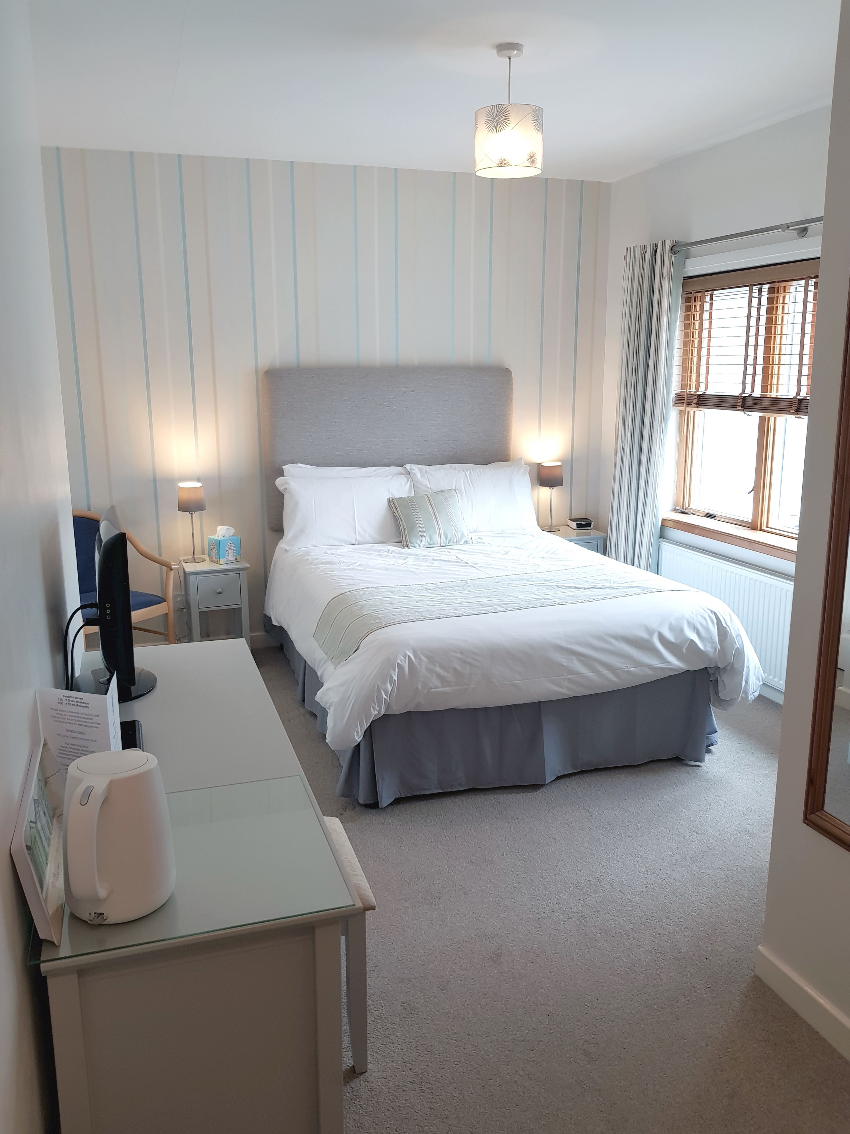 A neatly arranged bedroom with a bed, bedside lamps, a desk with a computer, and a window with curtains.