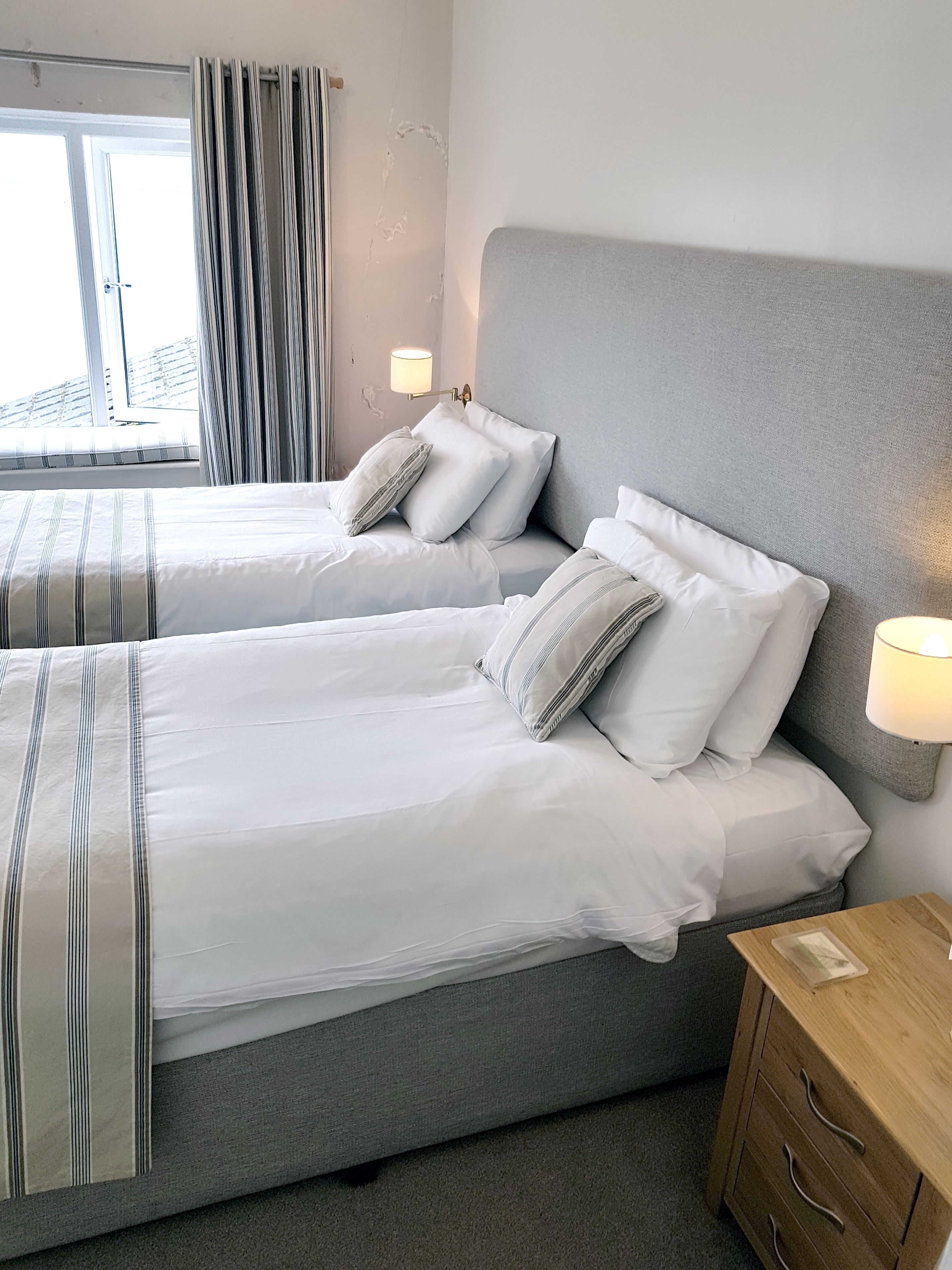A hotel room with two beds, a grey headboard, a bedside table with a lamp, and a view of the outside.
