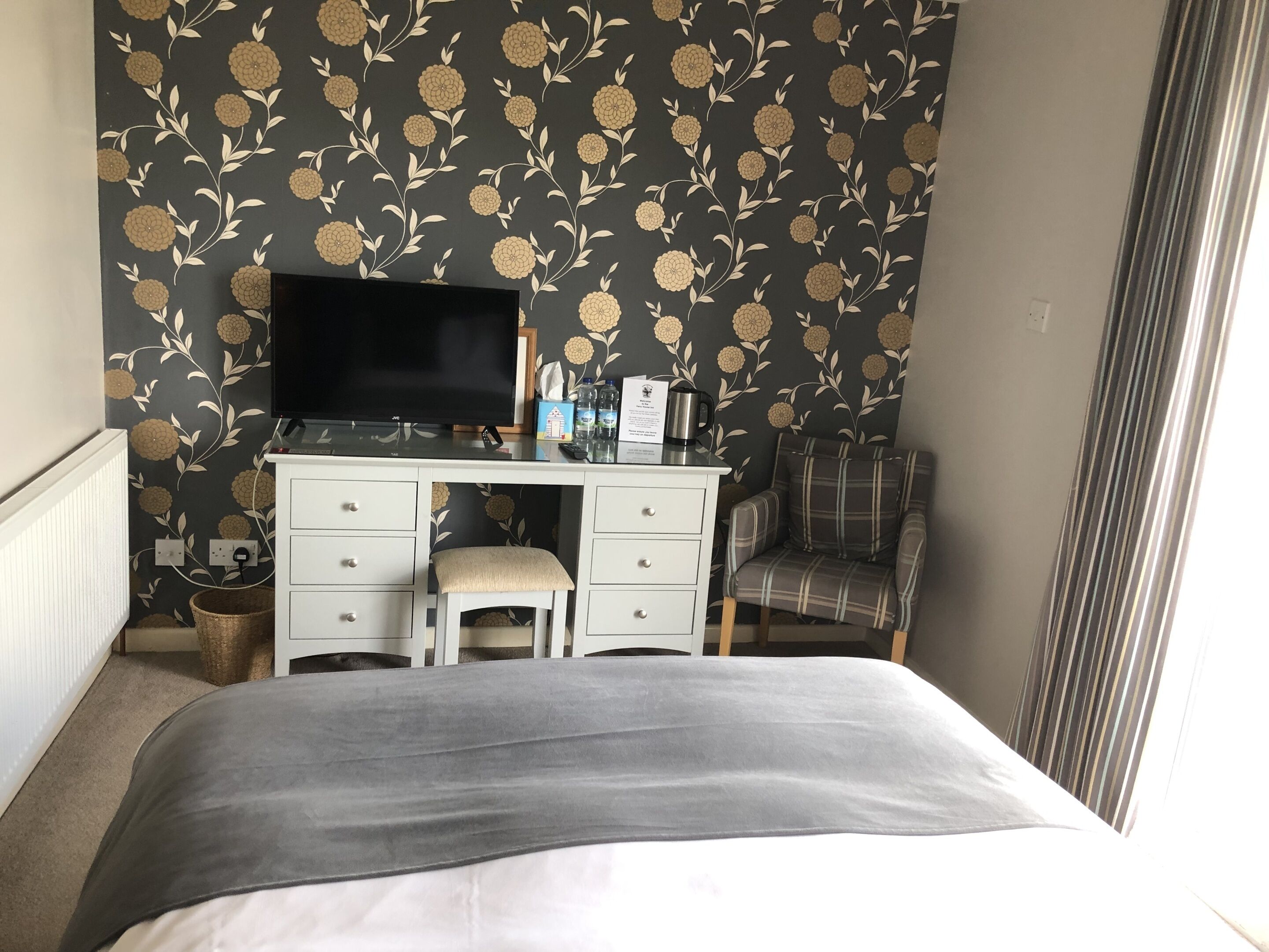 A bedroom with a bed, a desk with a TV, a chair, and a wall with floral wallpaper.