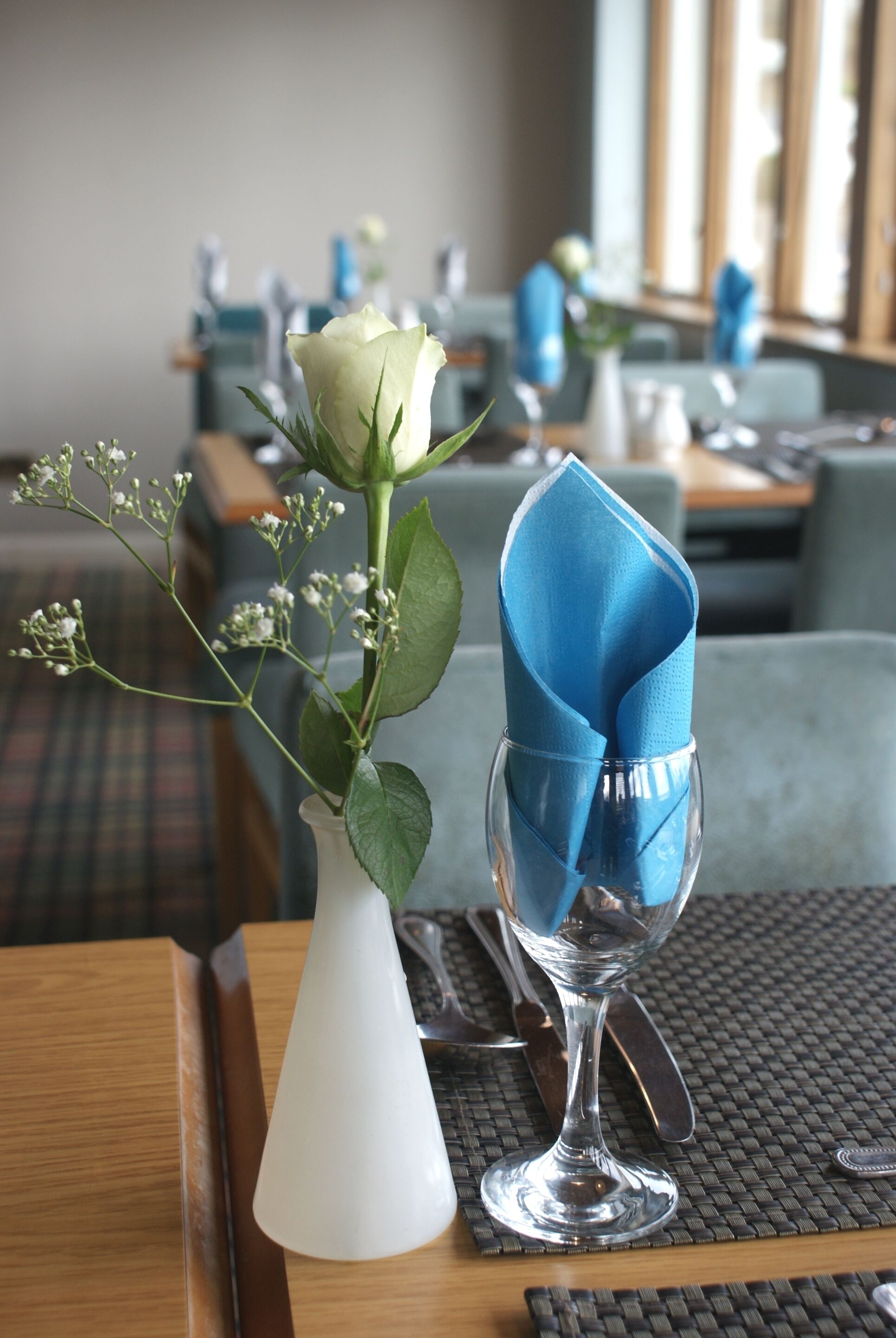 Une table à manger avec un vase blanc contenant une rose blanche et du gypsophile, une serviette bleue pliée et un verre à vin transparent.