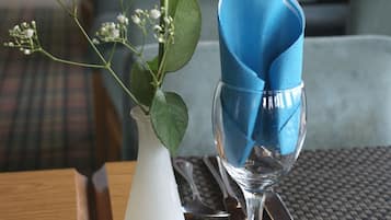 Une table à manger avec un vase blanc contenant une rose blanche et du gypsophile, une serviette bleue pliée et un verre à vin transparent.