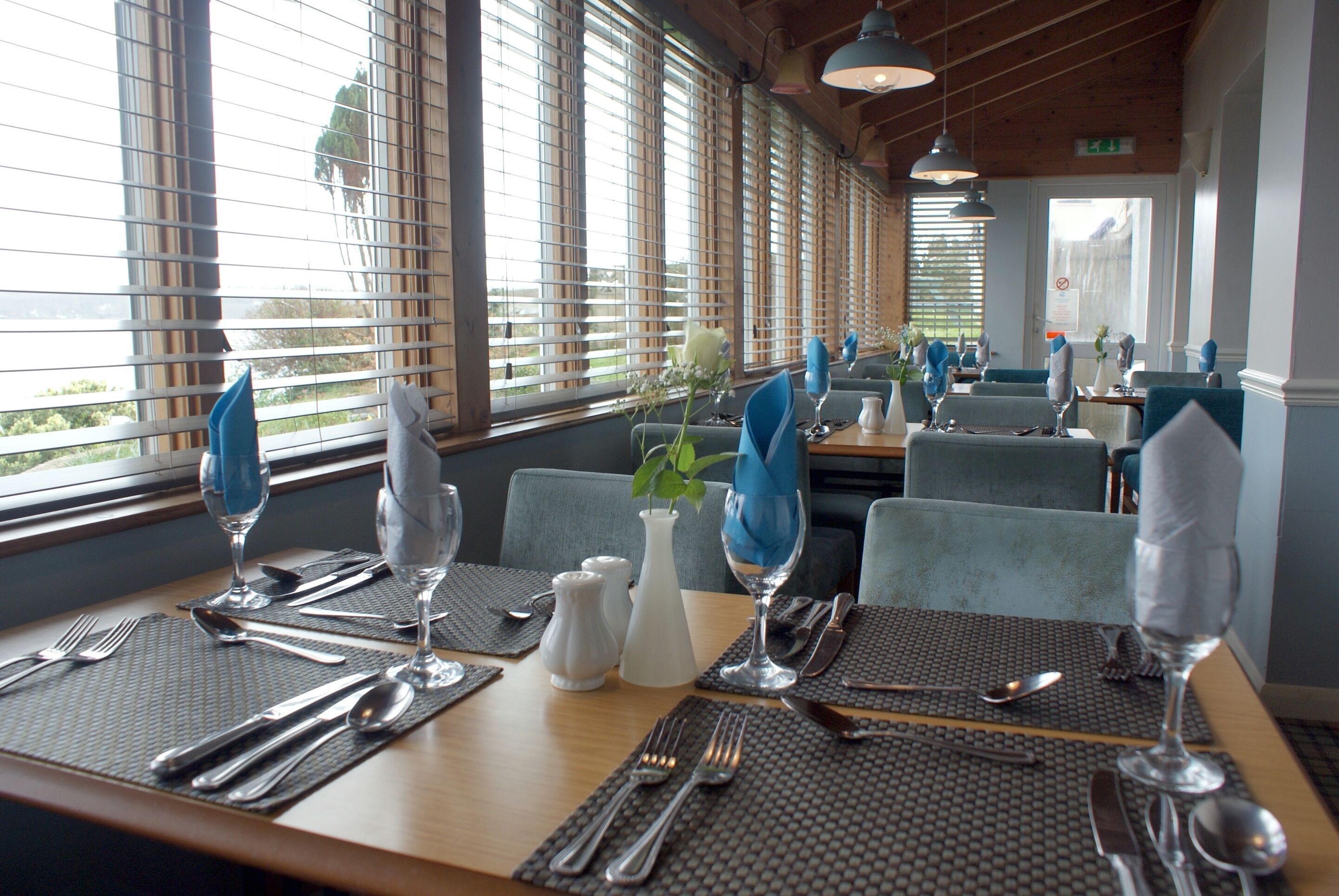 A restaurant with tables set for dining, featuring blue napkins, white salt and pepper shakers, and a vase with flowers.