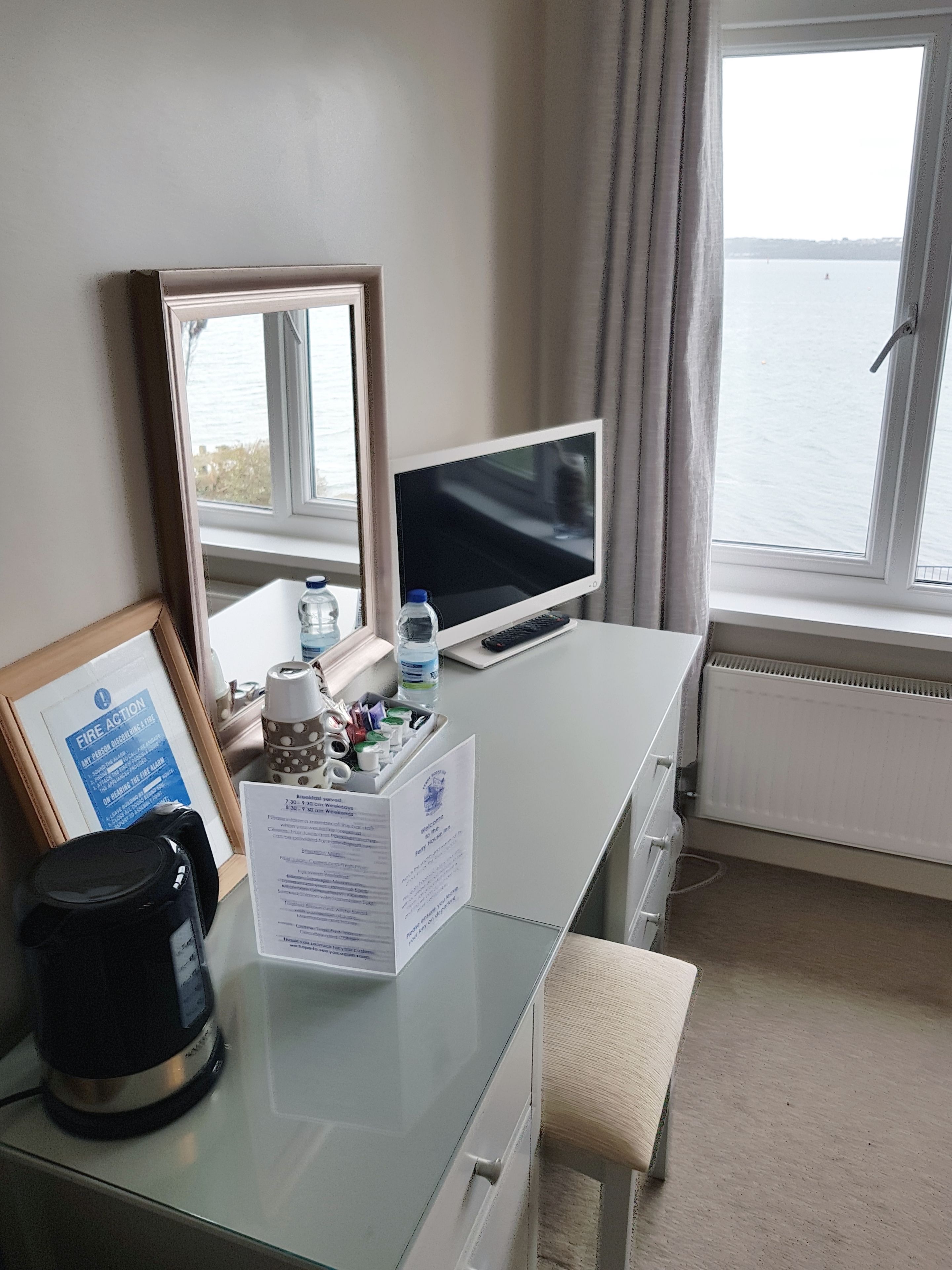 A hotel room with a desk, a television, a framed notice, a coffee maker, and a view of the sea through the window.