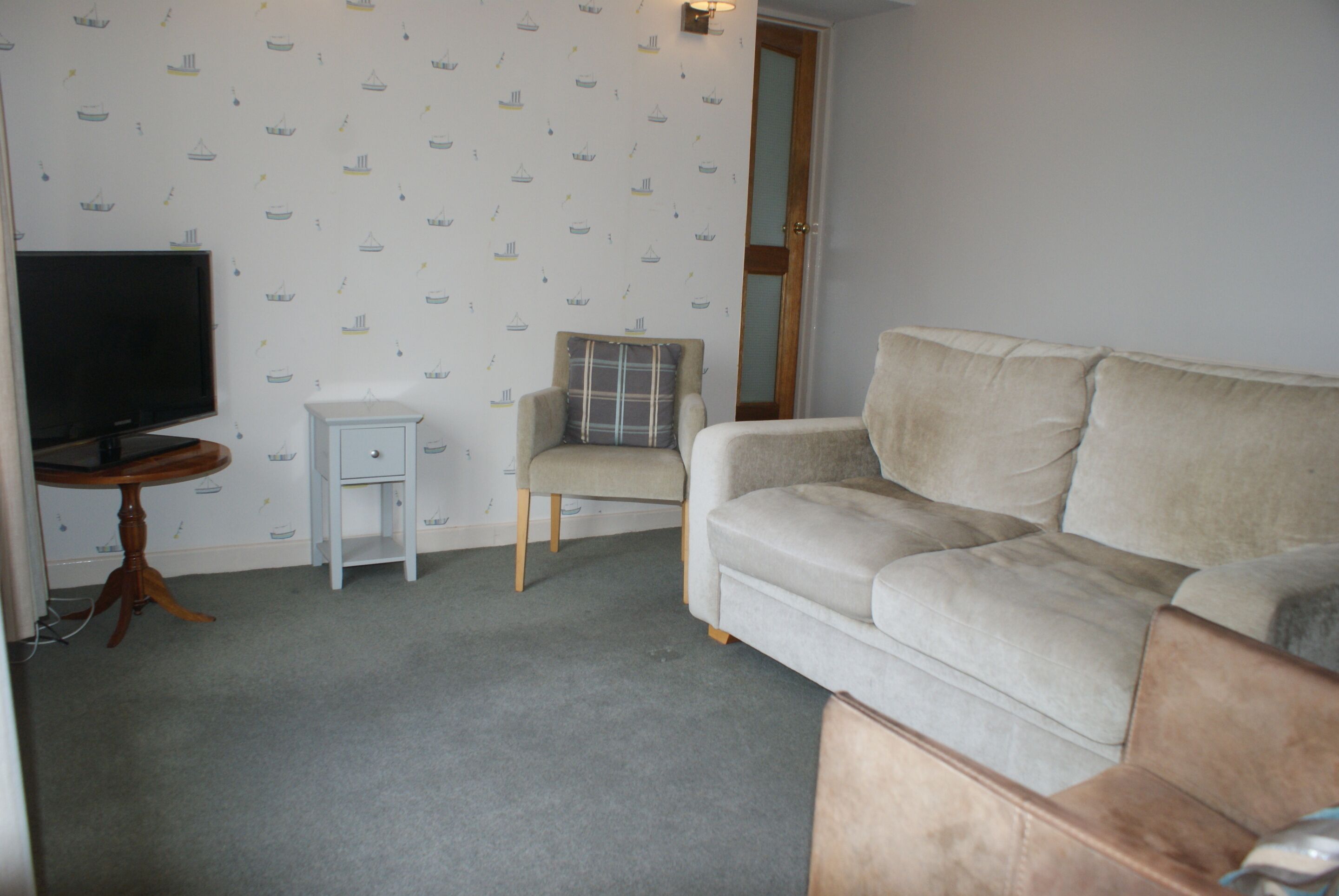 A living room with a beige sofa, a small white side table, a television, and a chair with a plaid cushion.