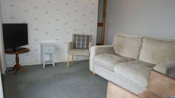 A living room with a beige sofa, a small white side table, a television, and a chair with a plaid cushion.