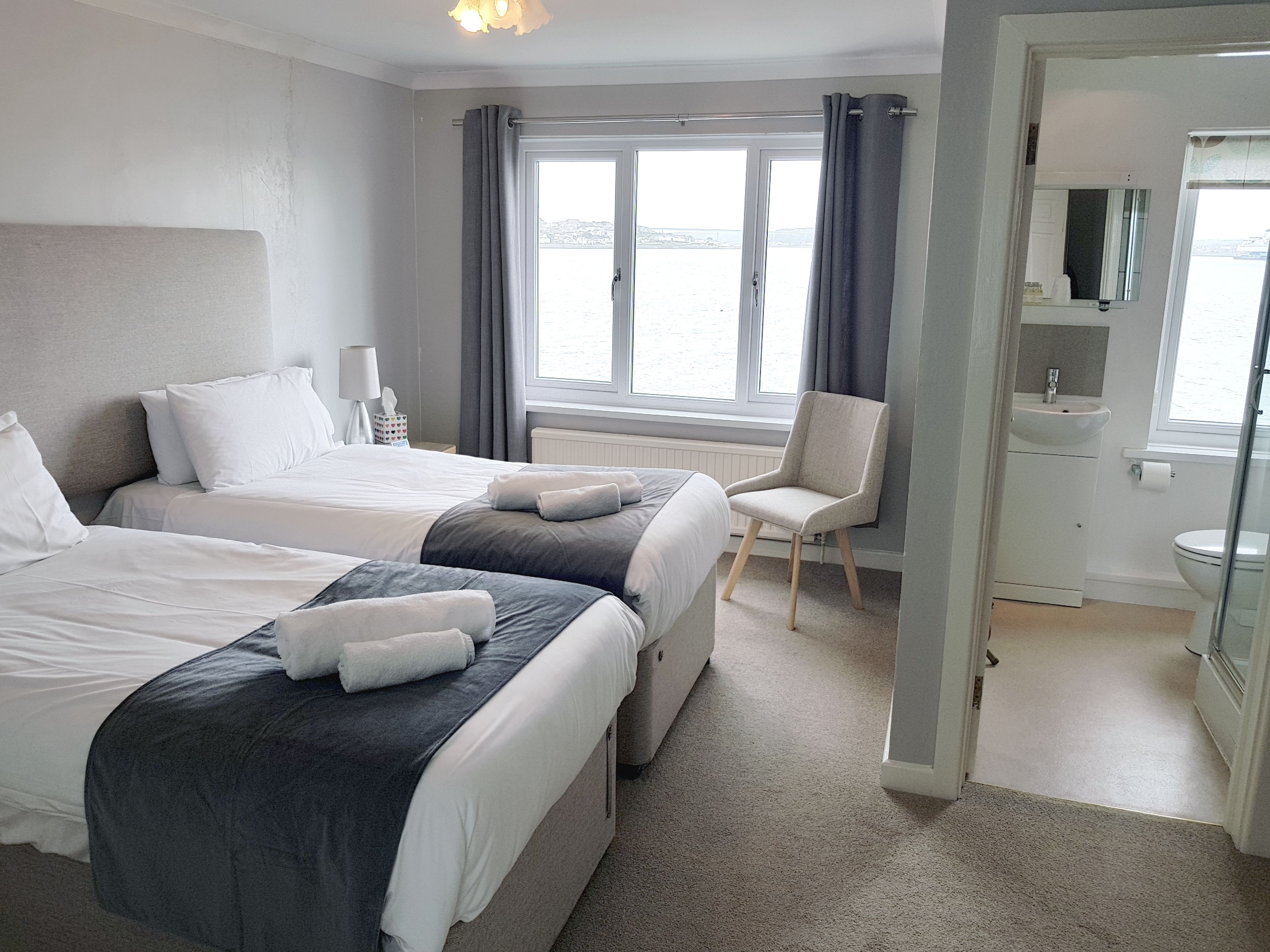 A hotel room with two beds, a chair, a bathroom with a sink and toilet, and a view of the sea through large windows.