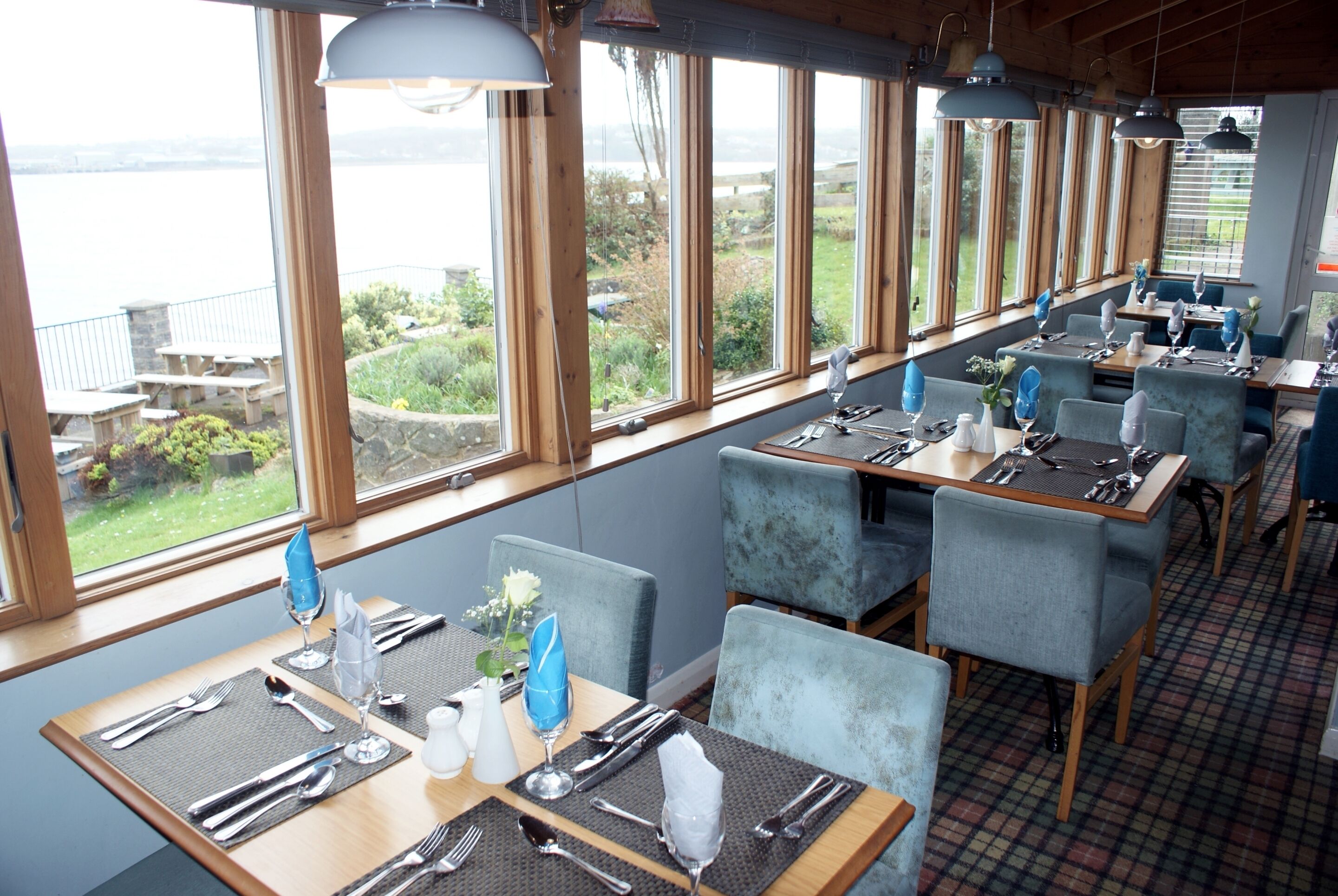 Un restaurant avec des tables dressées pour le service, de grandes fenêtres donnant sur l’extérieur et un plafond en bois.