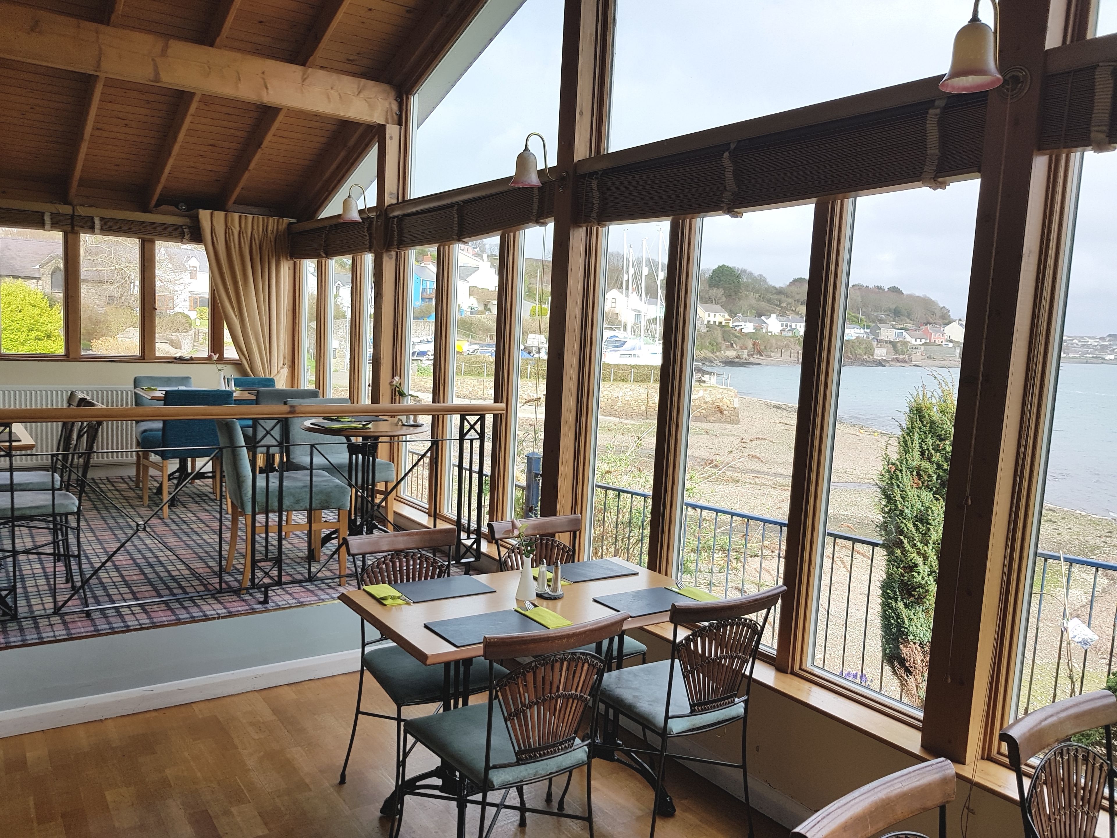 A restaurant with a view of the sea, featuring wooden tables and chairs, large windows, and a high ceiling.