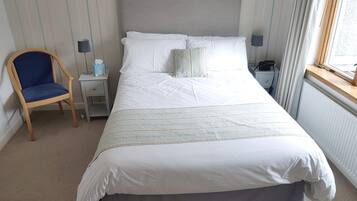 A neatly made bed with white linens and a grey skirt, a wooden chair, a bedside table with a lamp, and a nightstand with a phone.