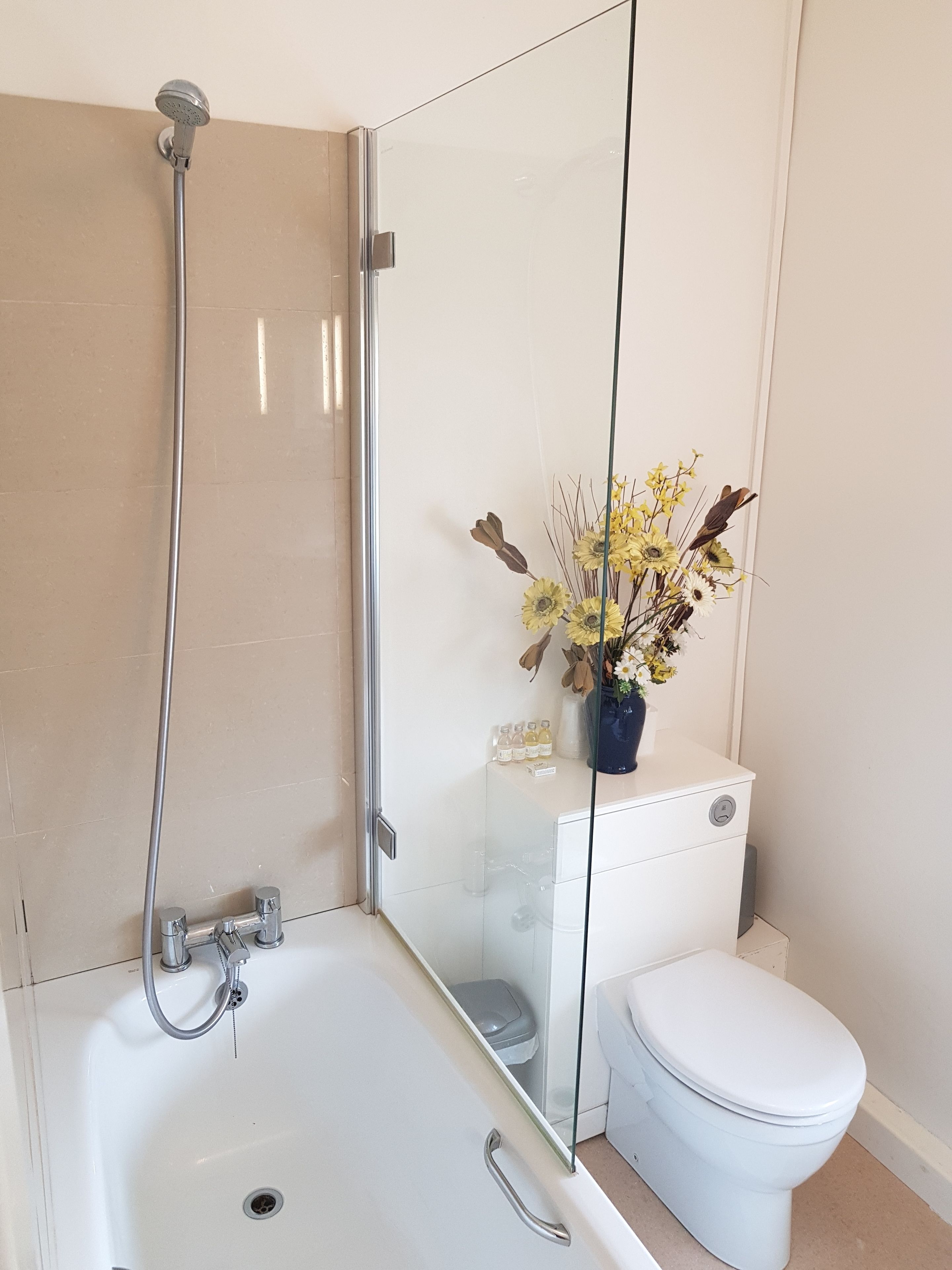Un baño moderno con cabina de ducha de vidrio, una bañera blanca y un inodoro con un jarrón azul de flores secas en la pared.
