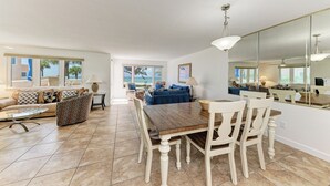 Un salón espacioso con una mesa de comedor, sillas y un sofá. Hay un espejo grande en la pared y vistas al exterior a través de las ventanas.