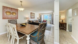 A dining area with a wooden table and white chairs, a wall with shell artwork, and a view through a doorway to a living room. - Unit #203 Sand Cay Beach Resort Gulf Front (Longboat Key)
