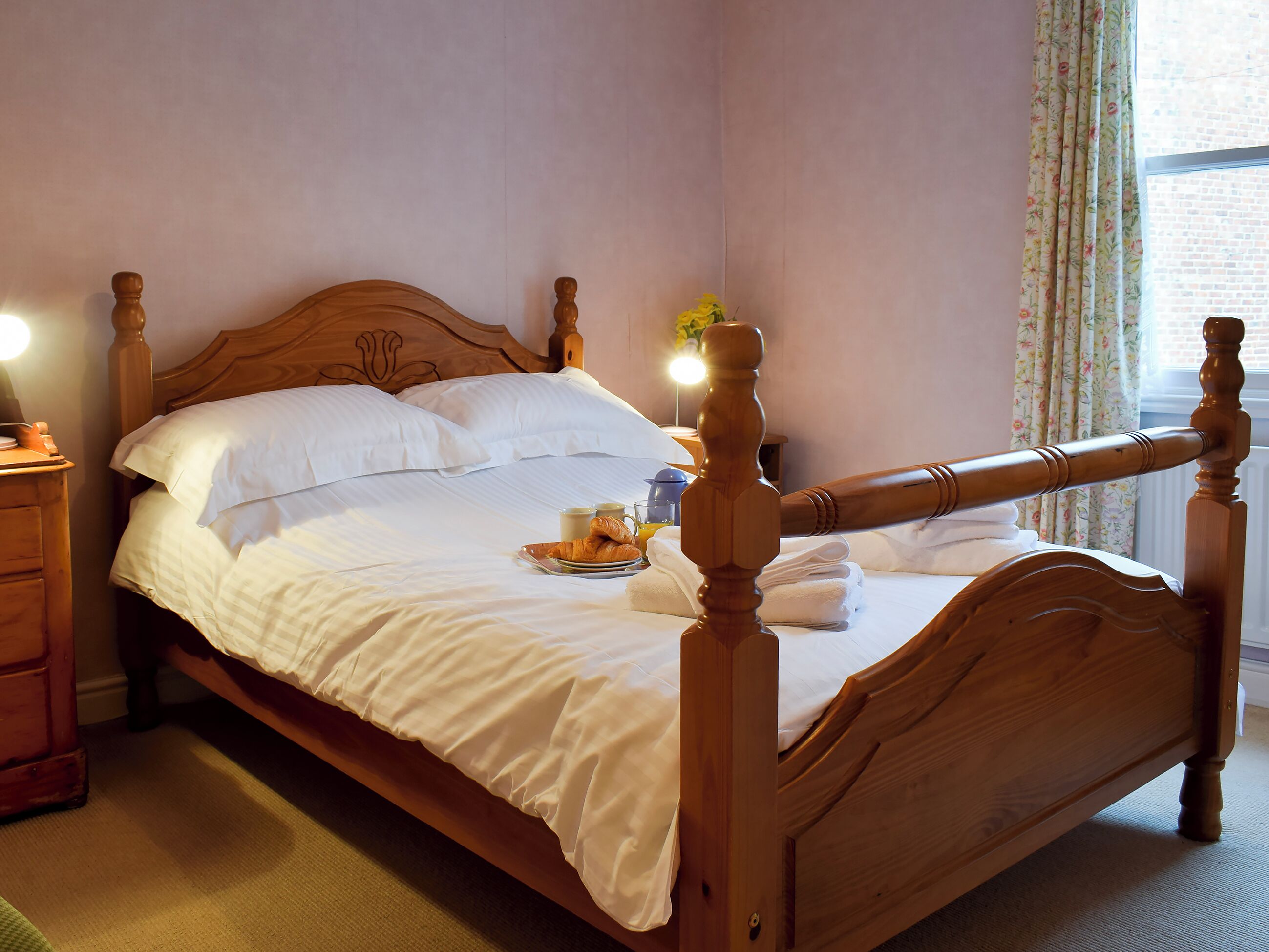 Ein hölzernes Himmelbett mit weißen Leinentüchern, ein Nachttisch mit Lampe und ein Fenster mit blumigen Vorhängen.