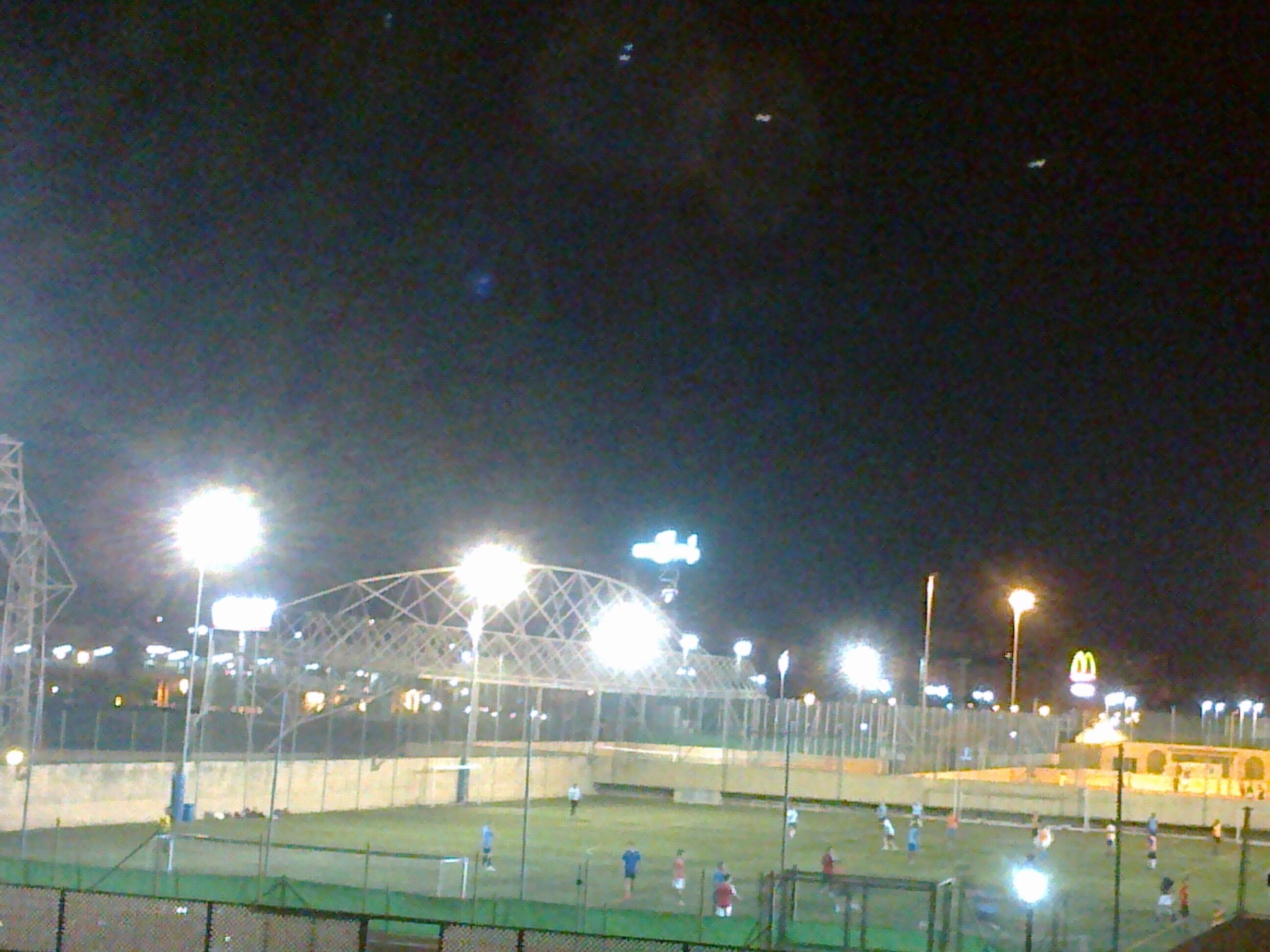 Vista nocturna de un campo deportivo con luces artificiales, una valla y un edificio distante con un letrero.