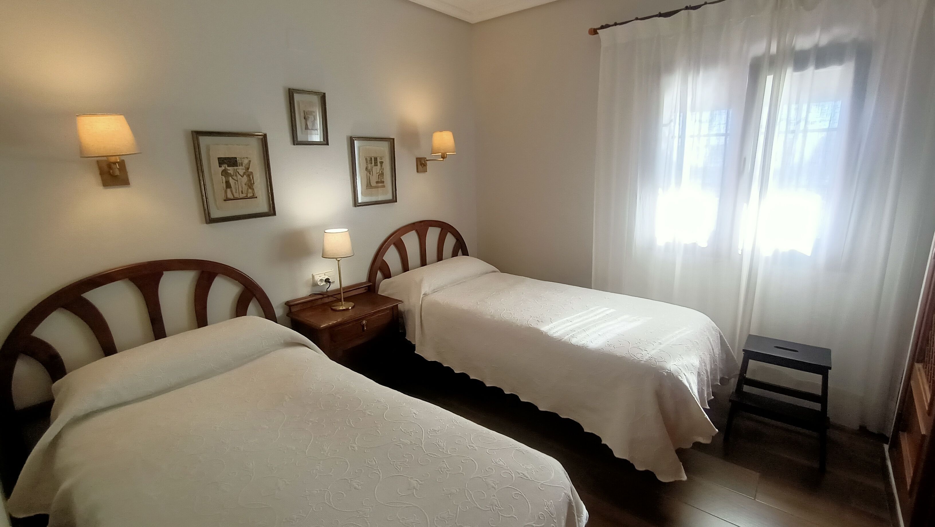 Une chambre d’hôtel avec deux lits, une table de chevet, des lampes fixées au mur, des tableaux encadrés, une fenêtre avec des rideaux légers et un petit tabouret en bois.