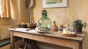 A wooden table with various food items and kitchenware, a green glass bottle, a painting, and a window with curtains. - The Old Priory Cottage (Dunster, near Minehead)