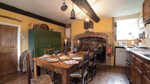 A rustic kitchen with a wooden table set for dining, a stone fireplace, and wooden cabinets. - The Old Priory Cottage (Dunster, near Minehead)