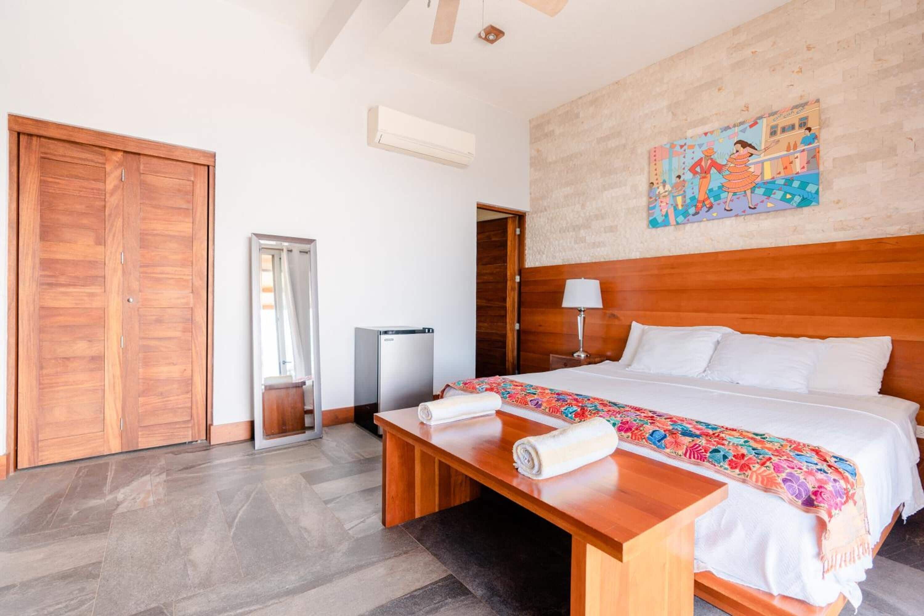Une chambre d’hôtel avec un lit en bois, un banc, une petite table, une poubelle et une armoire.