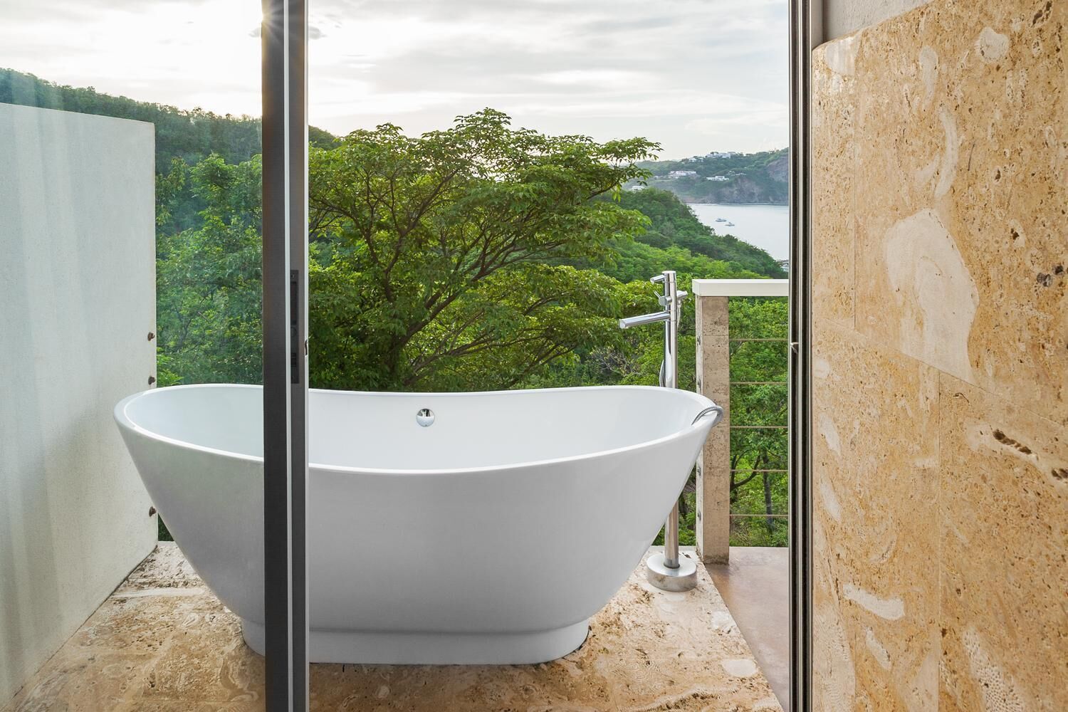 A freestanding bathtub in a balcony with a scenic view of a forest and water body.