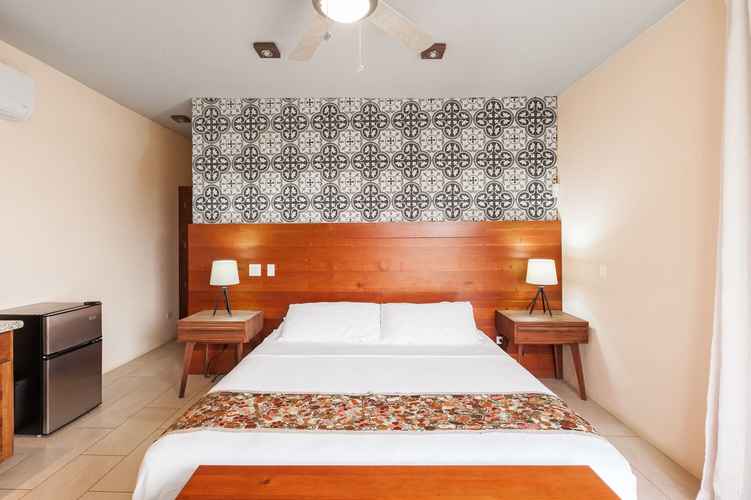 A hotel room with a wooden headboard, a patterned wall, a bed with white linens, two bedside tables with lamps, and a small refrigerator.