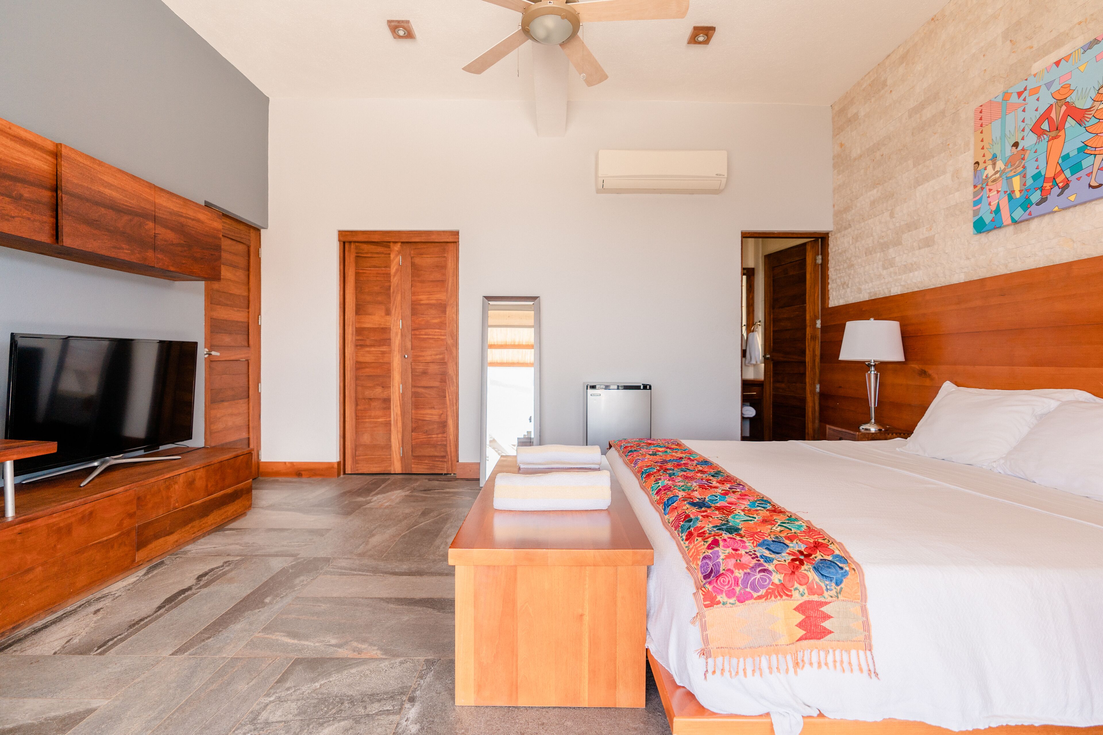 Une chambre d’hôtel avec un grand lit, une télévision, des boiseries, un ventilateur de plafond et une œuvre d’art colorée accrochée au mur.