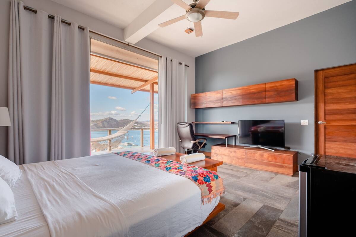Een hotelkamer met een bed, een bureau, een televisie en uitzicht op zee.