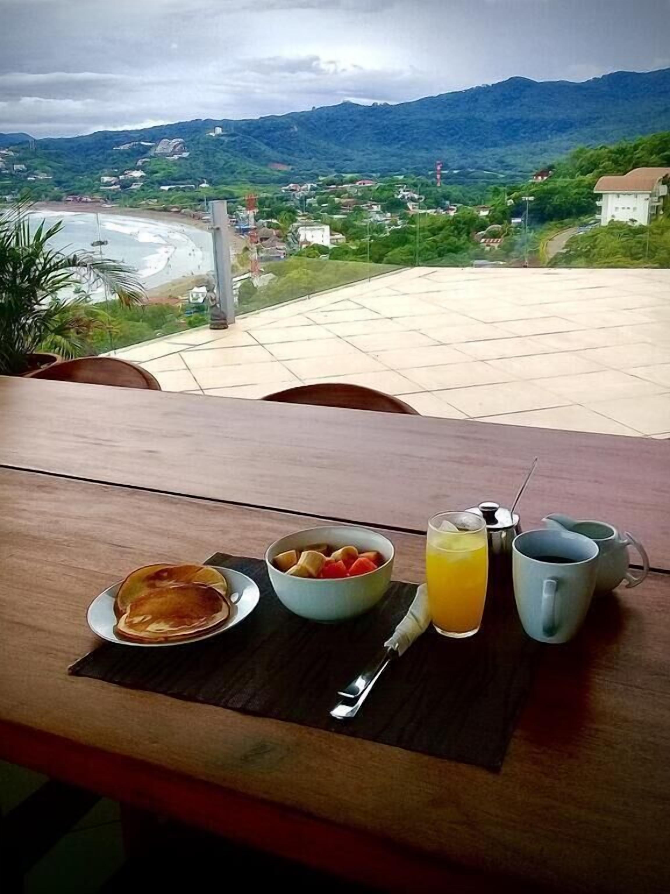 A table with breakfast items including pancakes, a bowl of fruit, a glass of orange juice, and two mugs, set against a scenic view of a coastal town and mountains.