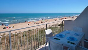 Un balcón con una mesa lista para comer, con vista a una playa donde hay gente y sombrillas.