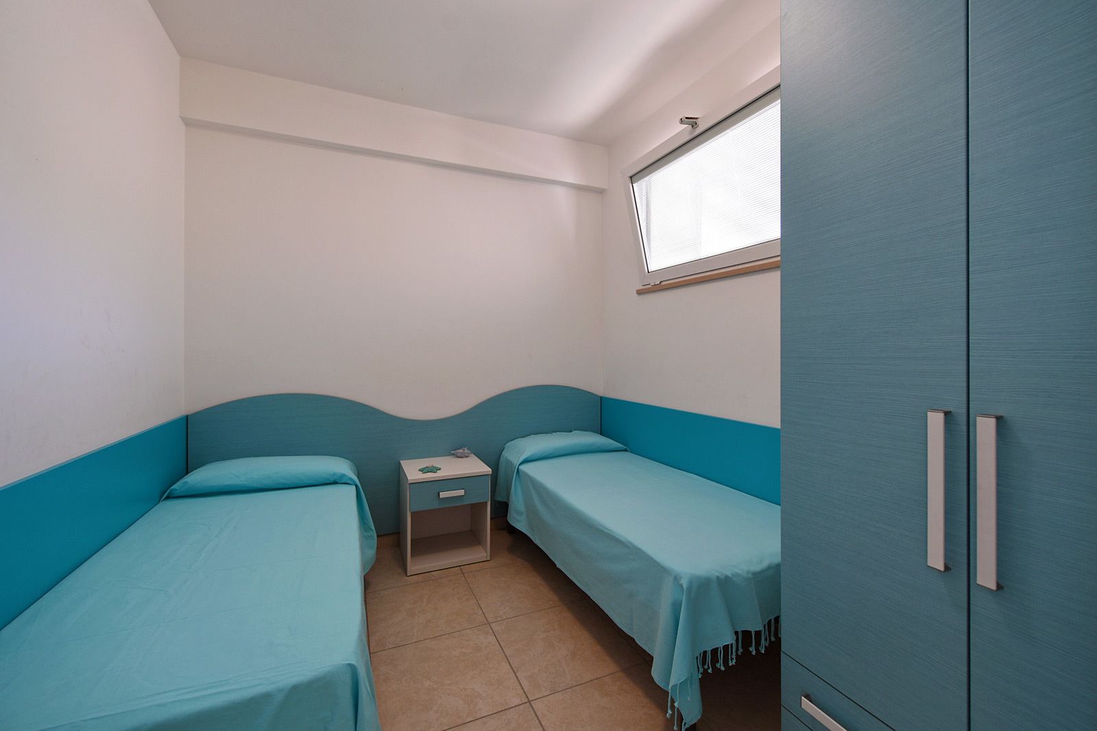 A small, single-room accommodation with two single beds, a small bedside table, a wardrobe, and a window with blinds.