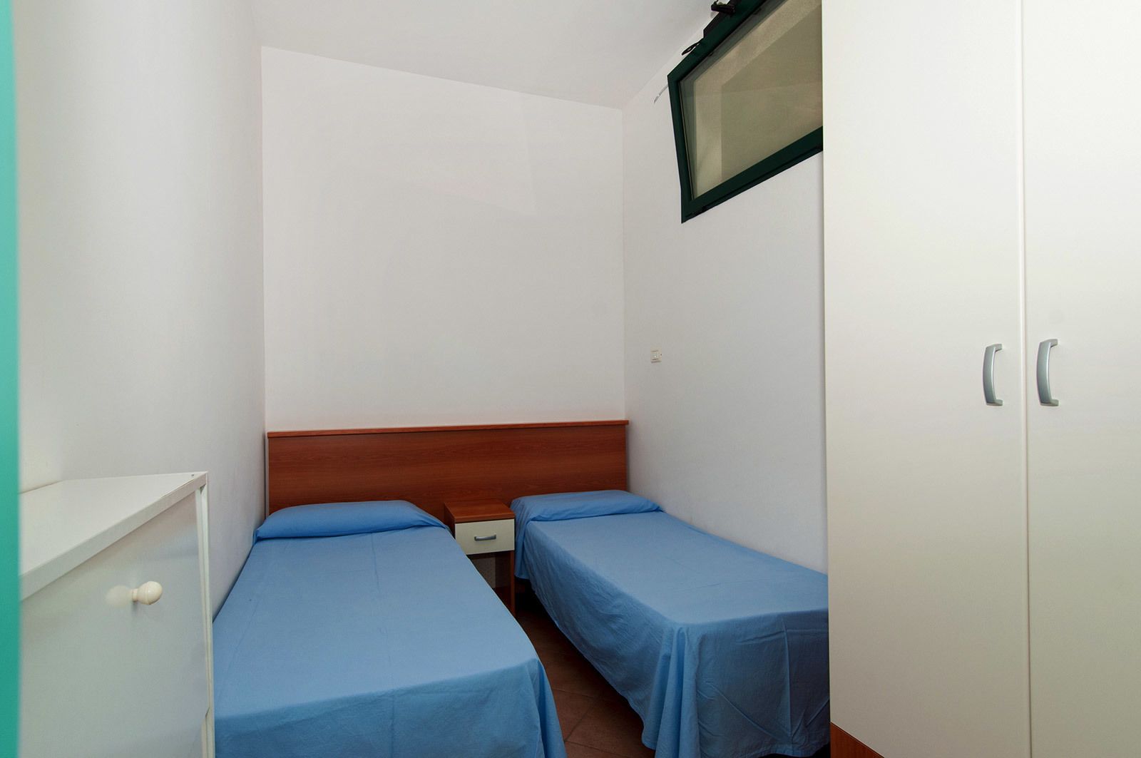 A small, simple room with two single beds, a wooden headboard, a white wardrobe, and a small window.