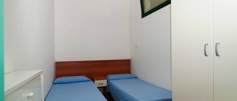 A small, simple room with two single beds, a wooden headboard, a white wardrobe, and a small window.