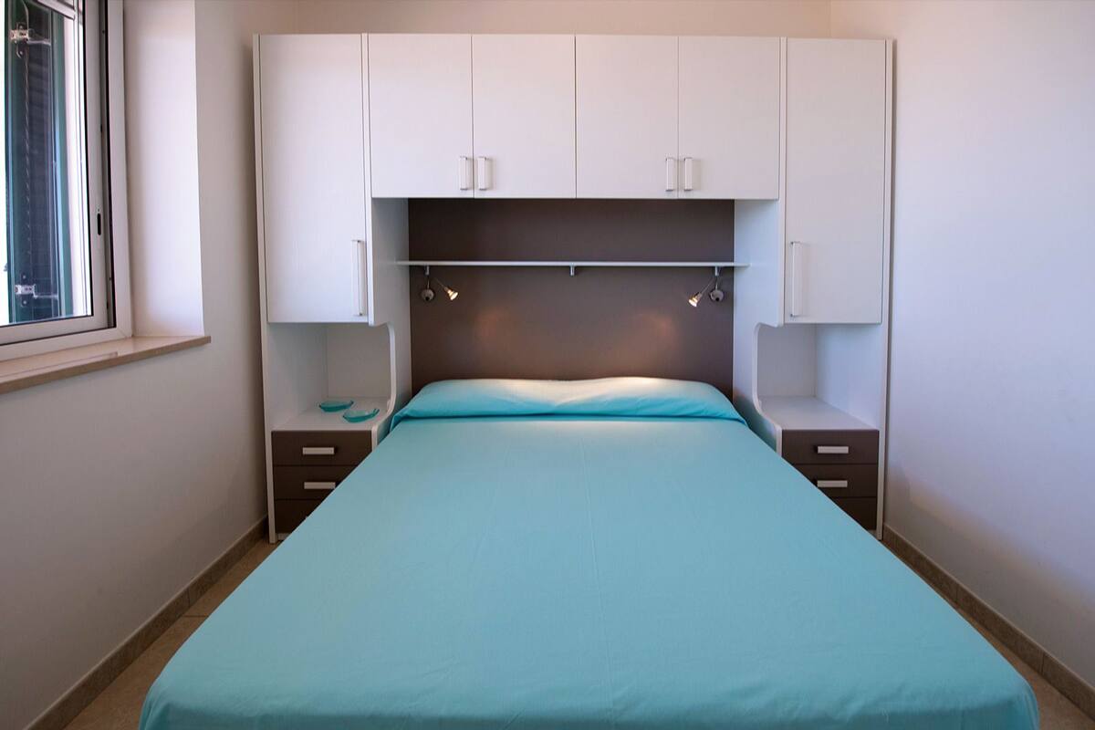 A bedroom with a bed, a nightstand, a drawer, a window, and a wardrobe.