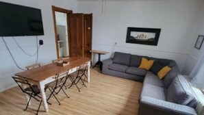 A living room with a wooden floor, a grey sofa with cushions, a wooden dining table with chairs, a ceiling fan, and a flat-screen TV mounted on the wall. - Family Wellness Space on 4 acres (Kensington)