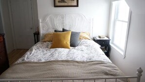 A neatly arranged bedroom with a bed, a bedside table, a nightstand, a framed picture on the wall, and a window providing natural light. - Family Wellness Space on 4 acres (Kensington)