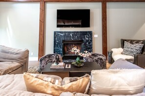 A living room with a fireplace, a flat-screen TV mounted above it, and a coffee table with books and a remote control.