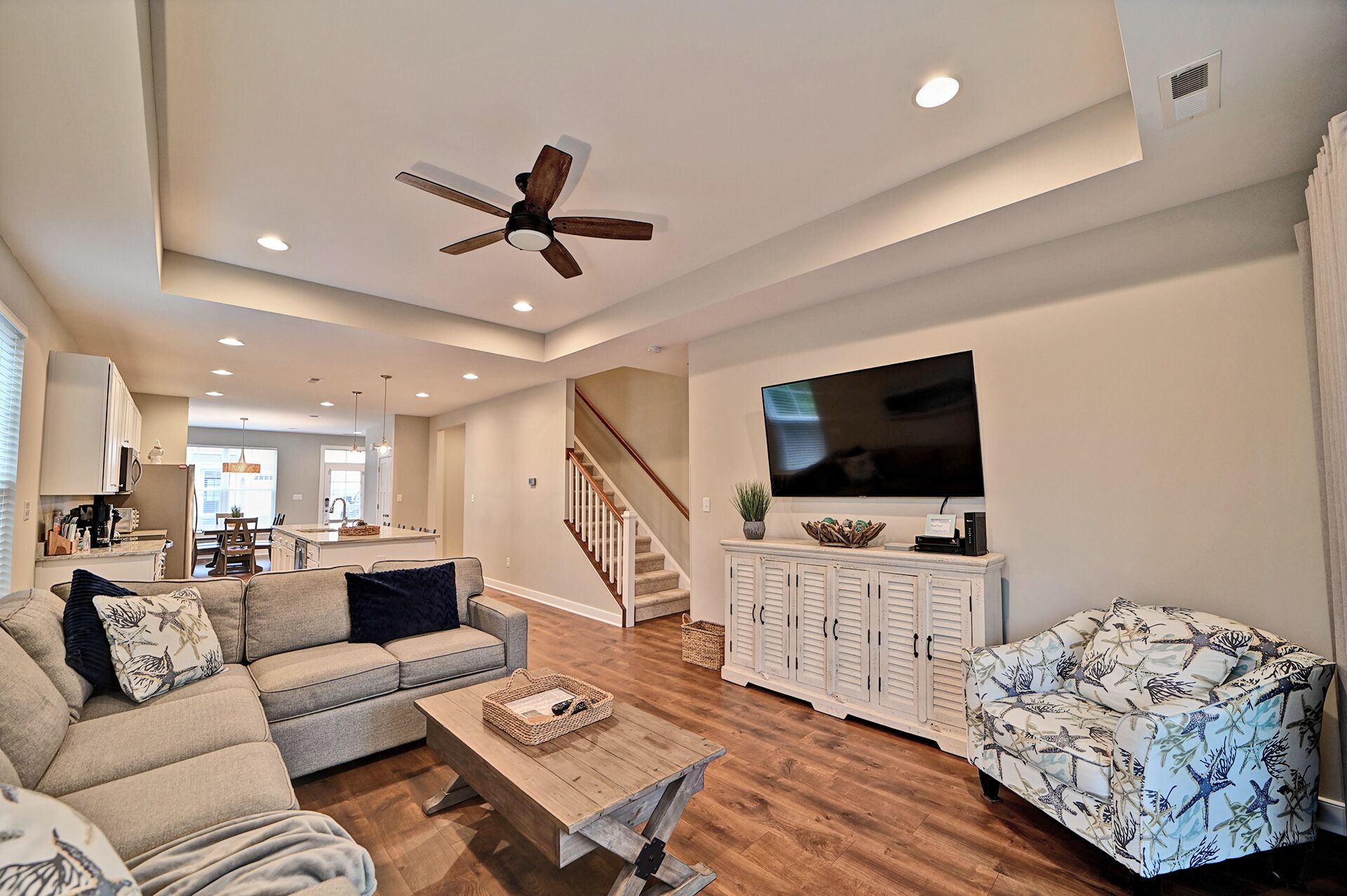 A spacious living room with a sofa, a coffee table, a patterned armchair, a ceiling fan, recessed lighting, a TV mounted on a cabinet, and a staircase leading to an open kitchen area.