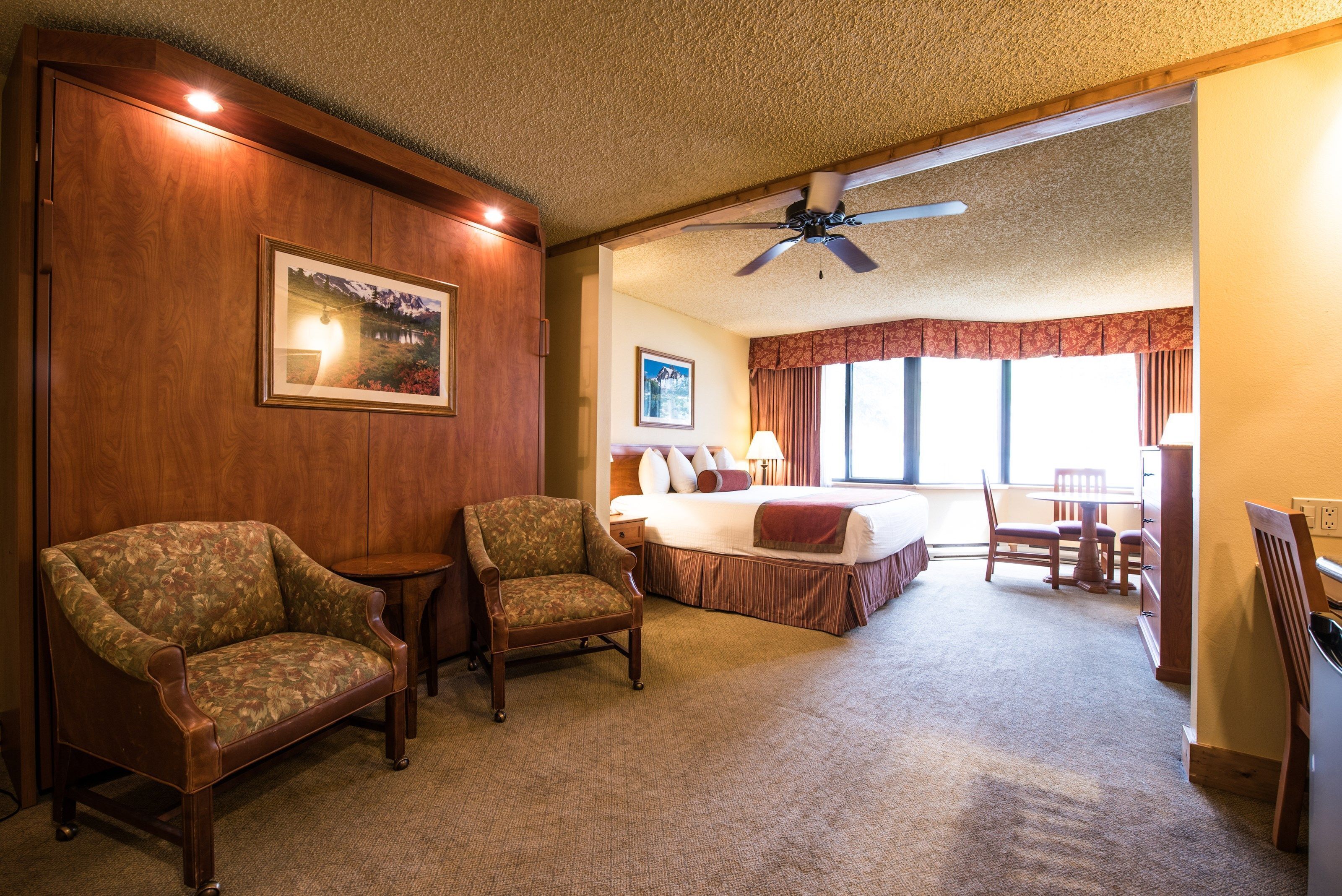 A hotel room with a bed, two armchairs, a desk, and a ceiling fan.