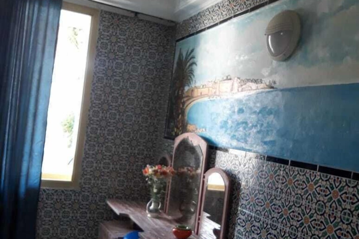 A room with patterned walls, a large mirror, a wooden dresser, and a window with curtains.