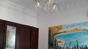 A bedroom with a bed, a nightstand, a wardrobe, a painting of a landscape, and a chandelier. - Hotel Hamilton - Kaly Center (Hammamet)