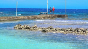 A clear blue sky, calm sea, rocky shoreline, and two people standing on a pier. - Reef House (Roatan)