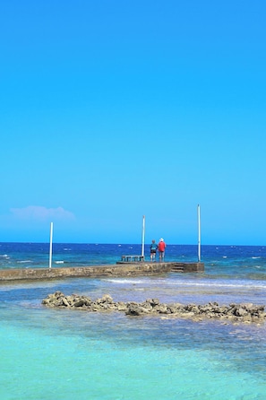 Cielo azul despejado, mar tranquilo, costa rocosa y dos personas en un muelle.