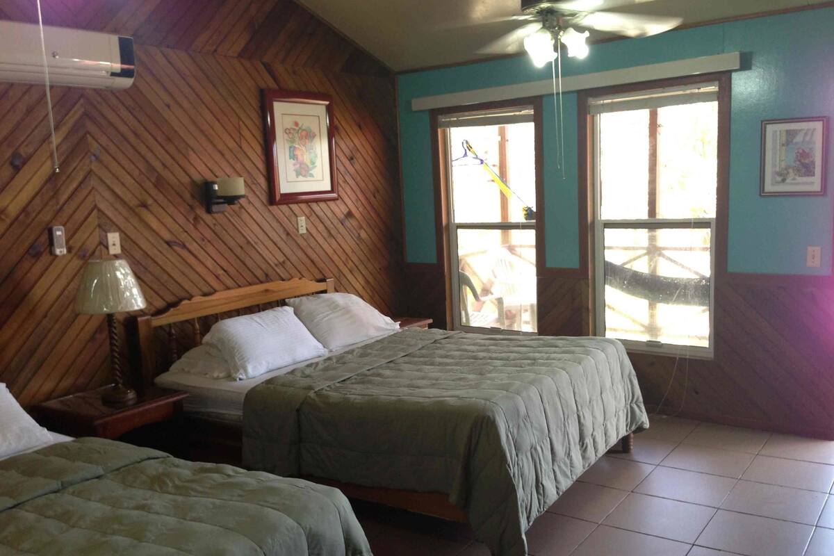 Una habitación con dos camas, ventilador de techo, paredes de madera y ventanas grandes.