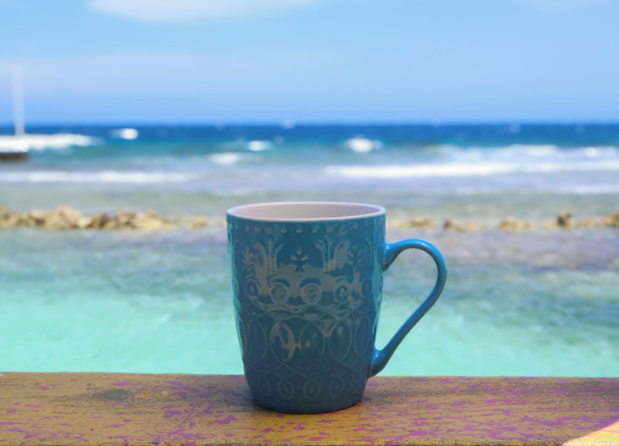 A single blue mug with a floral pattern on a wooden surface, overlooking a beach with clear water and distant boats.