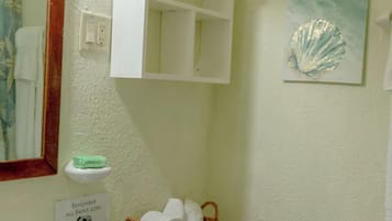 A bathroom with a white sink, a toilet, a basket of toilet paper, a shelf above the toilet, and a framed artwork on the wall.