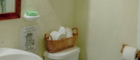 A bathroom with a white sink, a toilet, a basket of toilet paper, a shelf above the toilet, and a framed artwork on the wall.