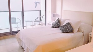 Et hotelværelse med en stor seng, to mønstrede puder, et natbord, en lampe og udsigt til den udendørs balkon med stole.