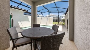 Un patio cubierto con una mesa redonda y sillas de mimbre, con vistas a una piscina y una zona residencial.
