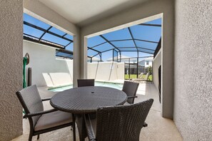 Un patio cubierto con una mesa redonda y sillas de mimbre, con vistas a una piscina y una zona residencial.