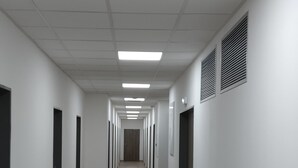 Un lungo e stretto corridoio con pareti bianche, pavimento in legno e luci fluorescenti al soffitto.
