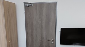 Una stanza con una porta in legno, una televisione fissata al muro, un tavolo da pranzo con un sottopiattino e un telecomando sul tavolo.